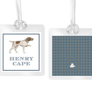 May include: Two square luggage tags with clear plastic loops. The left tag features a brown and white spotted dog illustration above the blue text HENRY CAPE. The right tag displays a blue and tan plaid pattern with a small white logo at the bottom.