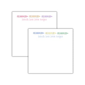 May include: Two white notepads with crayon illustrations and the text "lunch box love notes." One notepad features pink, orange, and purple crayons, while the other has blue, yellow, and green crayons. The notepads are designed for writing notes.