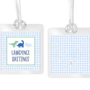 May include: Two square luggage tags with a light blue and white gingham pattern. One tag features three dinosaur silhouettes in green and blue, with the text LAWRENCE HASTINGS printed below. Both tags include clear plastic loops for attachment.