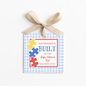 Puede incluir: Una etiqueta de regalo del Día de San Valentín con un patrón de cuadros vichy azul claro y una cinta beige. La etiqueta presenta gráficos de piezas de rompecabezas en amarillo, azul y rojo, con el texto "OUR FRIENDSHIP IS BUILT ON FUN! Happy Valentine's Day!"