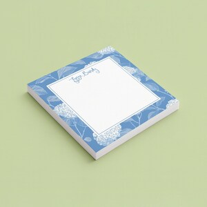 May include: A square notepad with a white center and a blue border featuring a white line-art floral pattern. The text "Linze Bandy" is printed in a blue script font at the top of the white area. The notepad has a stack of paper sheets.