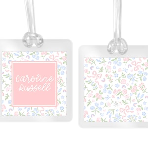 May include: Two square luggage tags with clear plastic frames and transparent loops. The tags feature a repeating pattern of small pink flowers, blue blossoms, green leaves, and pink bows. One tag displays the name Caroline Russell in white script.