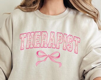 May include: A light beige sweatshirt with the word "THERAPIST" printed in pink letters with a pink bow underneath. The sweatshirt is being worn by a person sitting on a white couch.