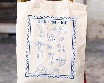 May include: Cream-colored tote bag with a blue stamp-like design featuring dog illustrations and the text "CHIEN + MOI = JOIE". The bag has two handles and is made of a sturdy fabric, perfect for carrying groceries or personal items.
