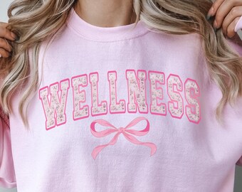 May include: Pink sweatshirt with the word "WELLNESS" in pink floral letters and a pink bow.