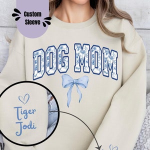 May include: A beige sweatshirt with the text "DOG MOM" in blue and white lettering with a blue floral pattern. The sweatshirt has a blue bow with a floral pattern. The text "Tiger Jodi" is on the sleeve in blue.