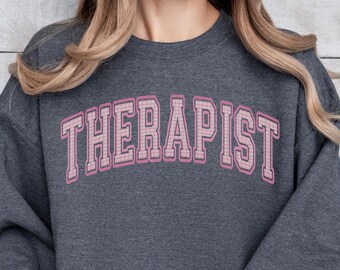 May include: A dark gray sweatshirt with the word "THERAPIST" printed in pink and white checkered letters.