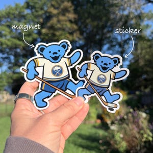 May include: A blue bear character wearing a cream and blue hockey jersey with a buffalo logo, holding a hockey stick. The image features two versions of this design, labeled as magnet and sticker, with text pointing to each item.