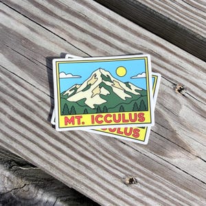 May include: A rectangular sticker featuring a graphic illustration of a snow-capped mountain peak under a light blue sky with a yellow sun and white clouds. Below the mountain, green pine trees are visible. The text MT. ICCULUS is printed in red.