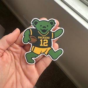 May include: A green bear sticker wearing a black and yellow football jersey with the number 12 and the word "Packers". The bear is holding a brown football. The sticker has a white border.