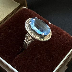 Könnte beinhalten: Ein Silberring mit einem ovalen blauen Edelstein, der in einen dekorativen Metallrahmen mit eingravierten Mustern gefasst ist. Der Ring befindet sich in einer braunen Schachtel mit dunkelrotem Samtinnenfutter und dem Aufdruck LDI, GERIA, ICERIA, RINO.