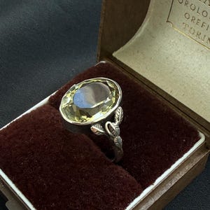 Puede incluir: Un anillo de plata con una gran piedra preciosa de color amarillo pálido y corte ovalado engastada en un bisel. La banda presenta un relieve detallado de pequeñas hojas que suben por los lados hacia el engaste, presentado en un joyero.