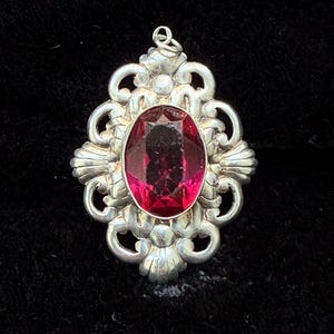 May include: A silver-toned pendant featuring an oval-shaped, deep red faceted gemstone set in the centre. The metal frame surrounding the stone displays an intricate, symmetrical scrollwork design with shell-like motifs at the top and bottom edges.