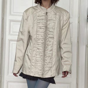 May include: A light beige jacket with a high collar and a central zipper closure. The jacket features vertical pleated detailing down the front and long sleeves. The jacket is worn over a black and white polka dot skirt and blue tights.