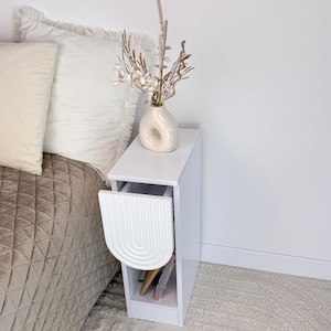 May include: A white bedside table with a decorative arched front. The table has a drawer and a top surface holding a vase with dried flowers. The table is next to a bed with pillows and a patterned comforter.