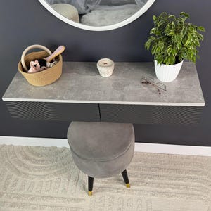 May include: A modern vanity set with a gray countertop and black drawers. A round mirror hangs above the vanity. Accessories include a woven basket, a candle, a potted plant, and a gray stool with black and gold legs.
