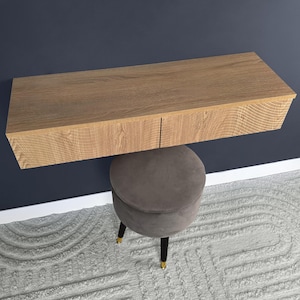 May include: A light brown, wall-mounted vanity with two drawers is positioned above a gray, velvet-covered stool. The stool has black legs with gold-colored tips. The background is a dark blue wall and a light gray patterned rug.