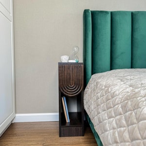 Può includere: Un comodino in legno scuro con un design curvo e uno scaffale per i libri. Il comodino è accanto a una testiera in velluto verde.