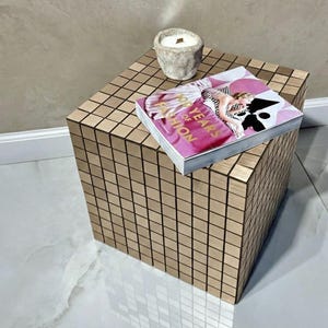 Op de afbeelding: Een vierkante, goudkleurige bijzettafel met een mozaïektegelontwerp. Een boek met de titel "100 Years of Fashion" en een kleine, steenkleurige kandelaar met een brandende kaars staan erop. De tafel staat op een wit en grijs betegelde vloer.