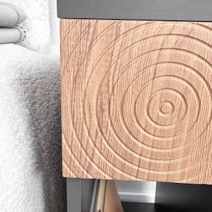 May include: Close-up of a wooden cabinet drawer with a concentric circle design. The drawer is made of light brown wood and has a dark gray top. The design is carved into the wood, creating a textured surface. The cabinet is part of a larger piece of furniture.