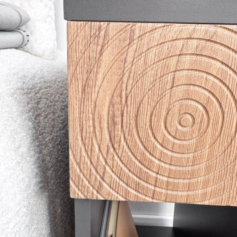 May include: Close-up of a wooden cabinet drawer with a concentric circle design. The drawer is made of light brown wood and has a dark gray top. The design is carved into the wood, creating a textured surface. The cabinet is part of a larger piece of furniture.