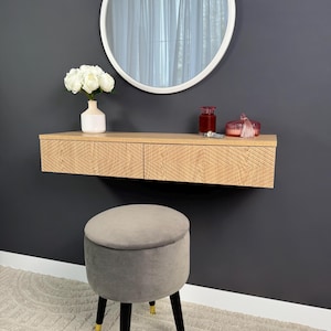 Peut inclure: Une coiffeuse en bois clair avec deux tiroirs et un motif géométrique. La coiffeuse est fixée à un mur gris foncé. Un miroir rond est suspendu au-dessus de la coiffeuse. Un vase blanc avec des fleurs blanches est posé sur la coiffeuse. Une petite bouteille en verre rouge et une boîte décorative rouge sont également sur la coiffeuse. Un tabouret rembourré gris avec des pieds noirs est placé devant la coiffeuse.