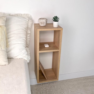May include: A light wood floating shelf with two shelves. The shelf is mounted to a white wall and has a small potted succulent plant, a small white object, and a book on the shelves.