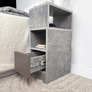May include: A gray, three-tiered nightstand with a drawer. The nightstand has a faux concrete finish and a geometric pattern on the drawer front. The drawer is partially open, revealing the metal drawer slides. Books are visible on one of the shelves.