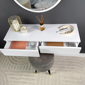Op de afbeelding: Een witte zwevende kaptafel met twee open lades, een grijze kruk en een ronde spiegel. De lades hebben een geometrisch patroon. Accessoires zijn onder meer kaarsen, sieraden en een geurverspreider. De kaptafel staat tegen een grijze muur.