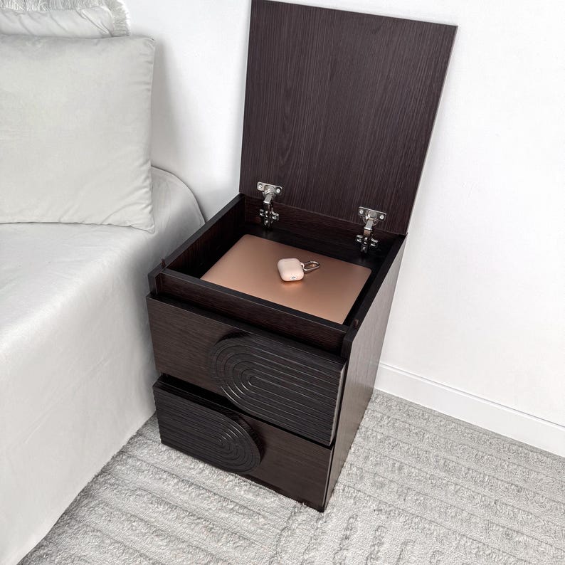 May include: A dark brown wooden nightstand with two drawers and a lift-top compartment. The top is open, revealing a rose gold laptop and a small, white wireless earbud case. The drawers have a decorative, semi-circular design.