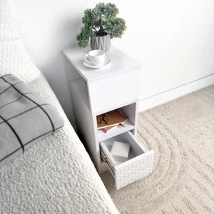 May include: A white bedside table with a drawer and open shelf. A small potted plant, a teacup and saucer, and books are on the table. The drawer has a textured, geometric design. The table is next to a bed with a gray blanket.