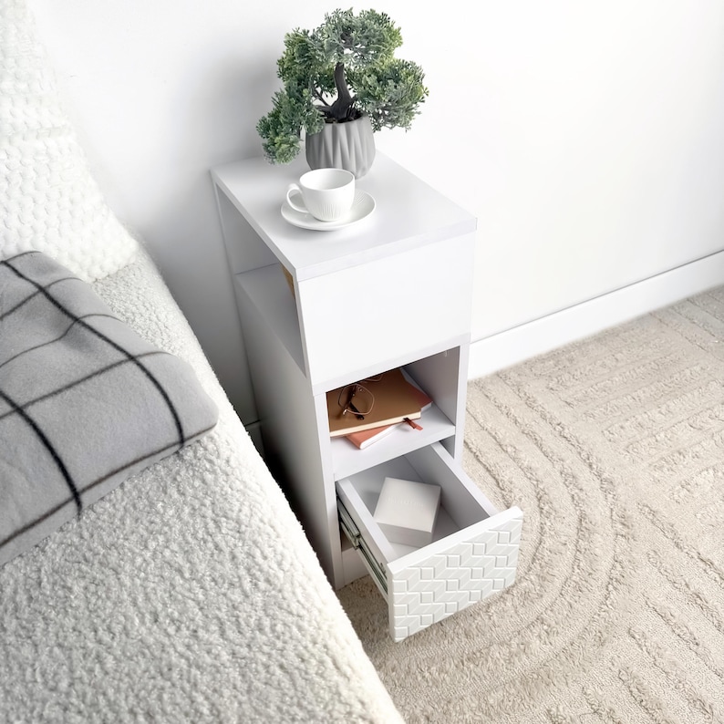 May include: A white bedside table with a drawer and open shelf. A small potted plant, a teacup and saucer, and books are on the table. The drawer has a textured, geometric design. The table is next to a bed with a gray blanket.