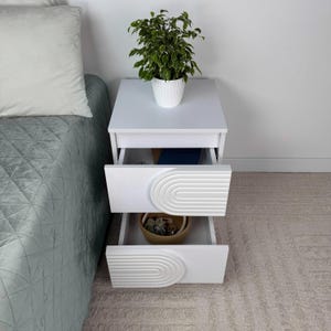 May include: White nightstand with two drawers, each featuring a decorative arched design. A potted plant sits atop the nightstand. The drawers are open, revealing contents. The nightstand is next to a bed with a green comforter.