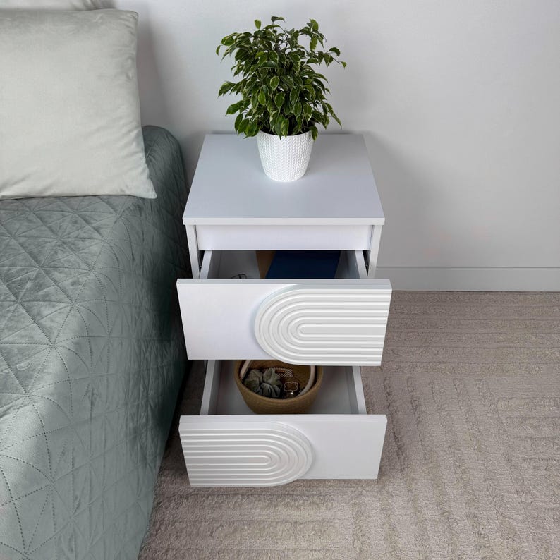 May include: White nightstand with two drawers, each featuring a decorative arched design. A potted plant sits atop the nightstand. The drawers are open, revealing contents. The nightstand is next to a bed with a green comforter.