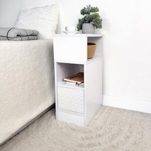 May include: A white bedside table with a geometric patterned drawer. The table has open shelving, a small woven basket, and a decorative plant. A cup and saucer sit on top. The table is next to a bed with a white comforter.