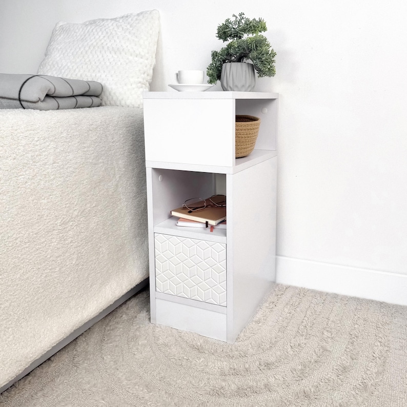 May include: A white bedside table with a geometric patterned drawer. The table has open shelving, a small woven basket, and a decorative plant. A cup and saucer sit on top. The table is next to a bed with a white comforter.