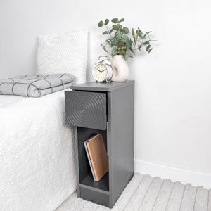 May include: A gray bedside table with a drawer featuring a concentric circle design. The open drawer reveals the interior, and a book is visible in the lower shelf. A white alarm clock and a vase with greenery sit on top.