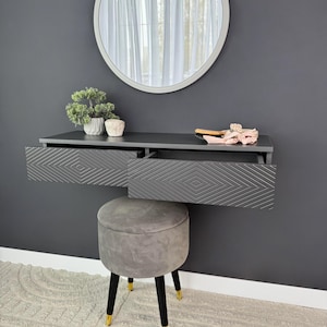 Puede incluir: Un tocador flotante negro con dos cajones que presentan un patrón geométrico. El tocador está montado en una pared gris con un espejo redondo encima. Un taburete gris con patas negras se encuentra delante del tocador.