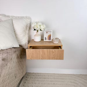 May include: A light wood floating nightstand with a single drawer. The drawer is open and has a wood grain pattern. The nightstand is decorated with a white vase with white flowers, a framed abstract print, and a small decorative object.