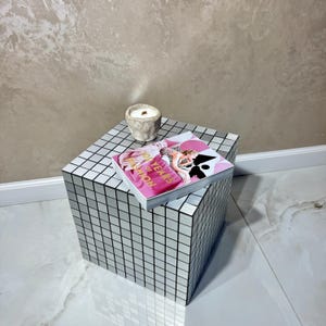 Op de afbeelding: Een spiegelende kubusvormige bijzettafel met een rasterpatroon, bekroond met een tijdschrift en een kleine kaars in een stenen pot. Het tijdschrift heeft een roze omslag met de tekst "100 Years of Fashion".