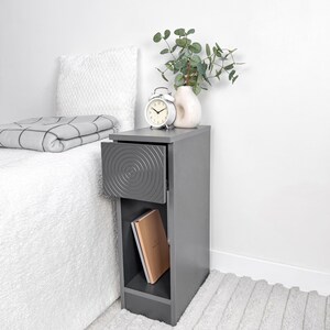 May include: A gray bedside table with a drawer featuring a concentric circle design. The drawer is open, revealing the interior. A white alarm clock, a vase with greenery, and a book are on the table. A folded blanket and pillow are on the bed.