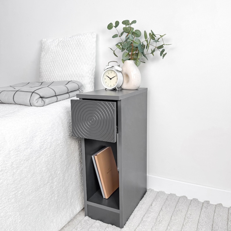 May include: A gray bedside table with a drawer featuring a concentric circle design. The drawer is open, revealing the interior. A white alarm clock, a vase with greenery, and a book are on the table. A folded blanket and pillow are on the bed.