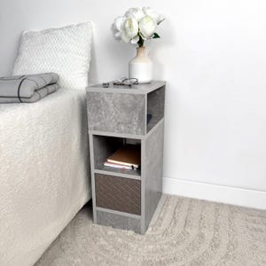 May include: A gray, modern nightstand with open shelving and a brown geometric patterned drawer. A white vase with white flowers, glasses, and notebooks are on top. The nightstand is next to a bed with a white comforter.