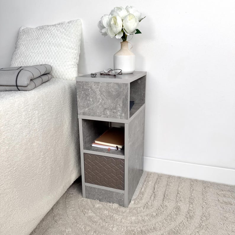 May include: A gray, modern nightstand with open shelving and a brown geometric patterned drawer. A white vase with white flowers, glasses, and notebooks are on top. The nightstand is next to a bed with a white comforter.