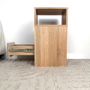 May include: A light brown wooden nightstand with a drawer that is partially open. The nightstand has a rectangular shape with a top open shelf. The drawer has metal hardware.