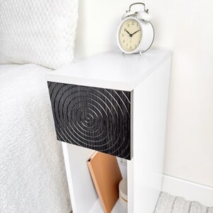 May include: A white bedside table with a black drawer front featuring a concentric circle design. A white alarm clock sits on top. A book and a container are visible in the open shelf below.