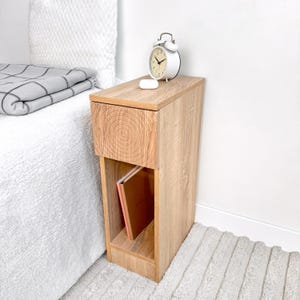 May include: A light brown wooden nightstand with a drawer and open shelf. A white alarm clock and small white object sit on top. A book is visible on the shelf. The nightstand is next to a bed with a white comforter and a gray blanket.