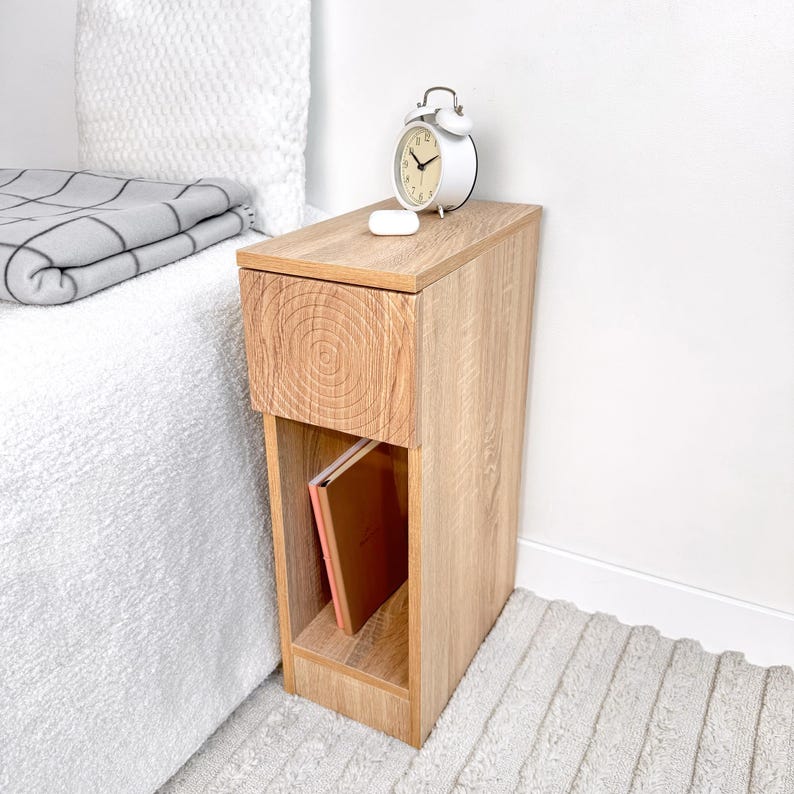 May include: A light brown wooden nightstand with a drawer and open shelf. A white alarm clock and small white object sit on top. A book is visible on the shelf. The nightstand is next to a bed with a white comforter and a gray blanket.