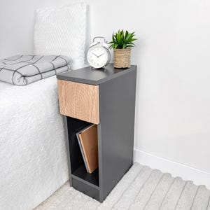 May include: A modern gray nightstand with a light wood drawer front featuring a circular grain pattern. A silver alarm clock and a small potted plant sit on top. A book is visible in the open shelf below.