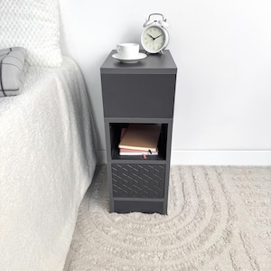 May include: A dark gray bedside table with a geometric patterned drawer. A white teacup and saucer sit on top, next to a white alarm clock. The table has an open shelf with books. The table is next to a bed with a white comforter.